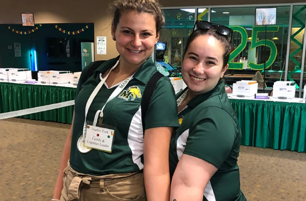 Two women wearing green and white polo shirts with name tags, smiling and standing in a decorated indoor setting.